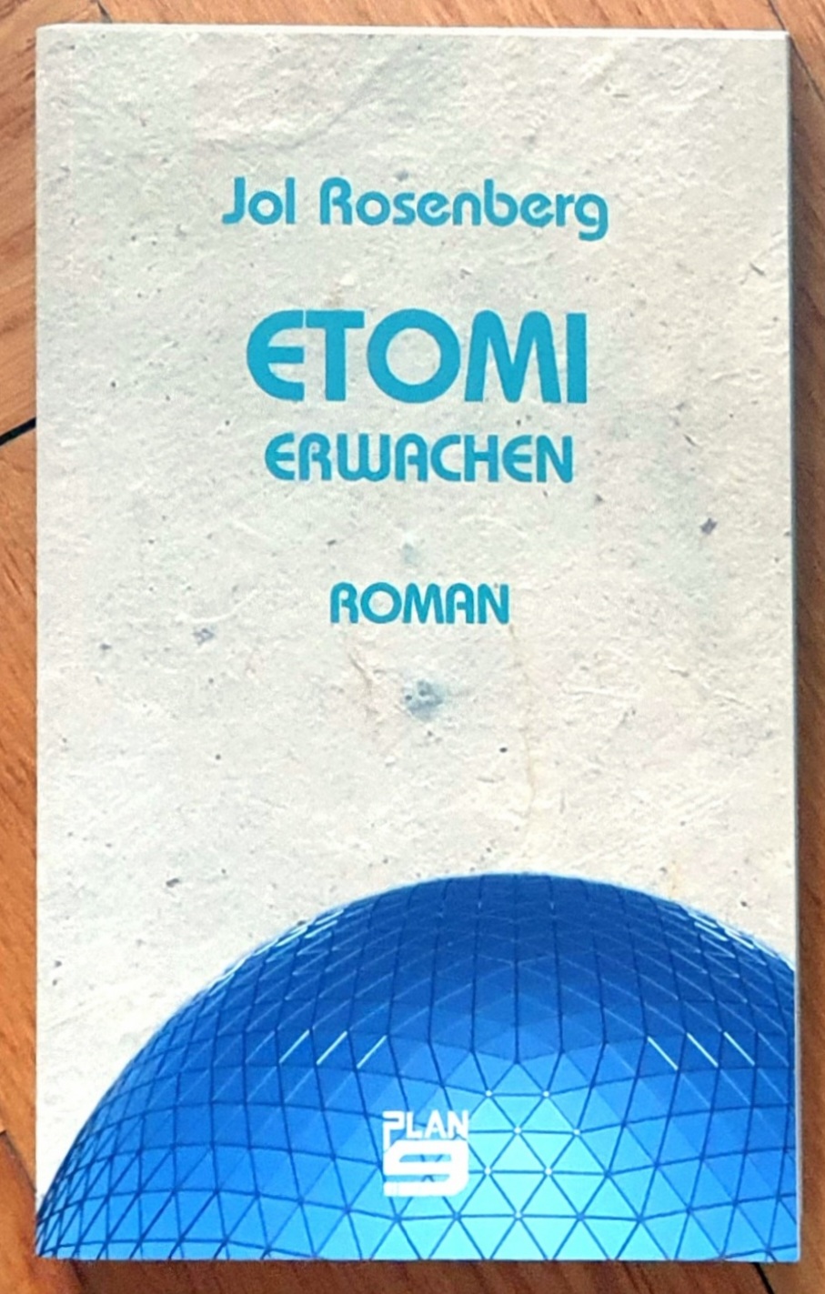 Buchdeckel von Etomi-Erwachen, von Jol Rosenberg bei Plan 9, blaue Schrift auf hellem Hintergrund wie selbstgeschöpftes Papier. Unten angeschnitten eine blaue Glaskuppel bestehend aus vielen dreieckigen Glasstücken die das Licht reflektieren.