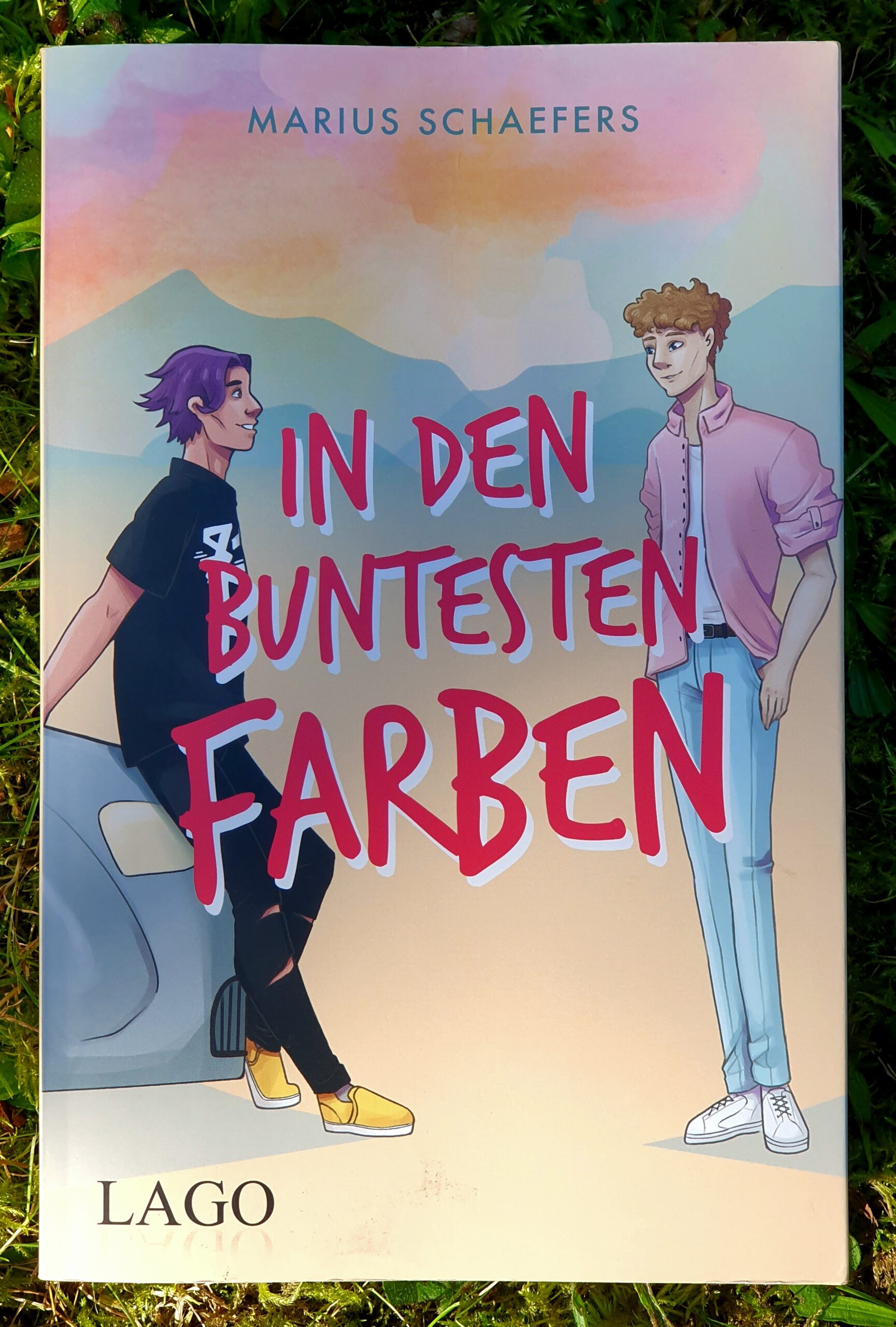 Einband des Romans: In den buntesten Farben von Marius Schaeffers bei LAGO. Der Autorenname und der Verlag jeweils am obere und unteren Seitenrand. In der Mitte zwei gezeichnete kolorierte Figuren. Einer mit lila Haaren und schwarzem Shirt lehnt an einem Auto, der andere mit braunen Locken und rosa Hemd, sie schauen sich an. Der Titel ist darüber in großen roten Buchstaben gedruckt.