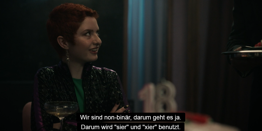 Screenshot mit Untertiteln aus der Serie: Glenda mit kurzen roten Haaren spricht zu einer anderen Person im Hintergrund: Wir sind non-binär, darum geht es ja. Darum wird 