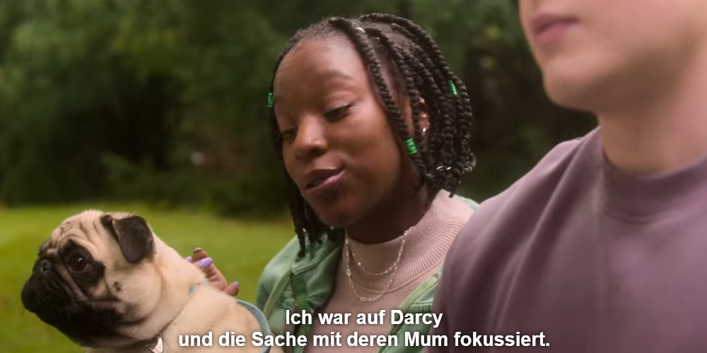 Screenshot aus der Serie. Ein schwarze Person mit halblangen geflochtenen Zöpfen mit Hund im Arm sitzt neben einer weißen Person, das Gesicht ist nicht richtig zu sehen. Untertitel: Ich war auf Darcy und die Sache mit deren Mum fokussiert.